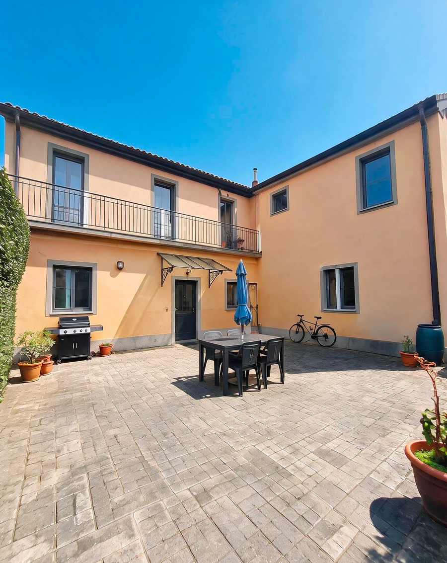 Cortile chiuso fra due ali della casa, con tavolo e ombrellone: la foto di partenza col cielo coperto e la pavimentazione bagnata, e la stessa inquadratura col sole