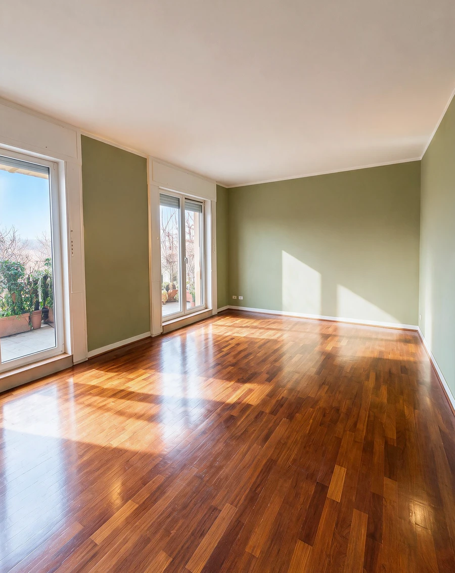 Soggiorno con pareti verde salvia e parquet scuro: la foto di partenza arredata e con la luce grigia, e la stessa stanza svuotata col sole che entra dalla portafinestra
